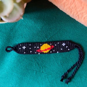 May include: Black friendship bracelet with a white and red planet design on a green background.