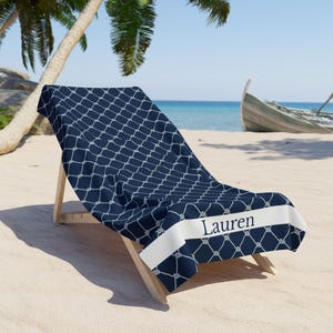 Puede incluir: Una toalla de playa azul marino con un patrón de red de cuerda blanca y el nombre "Lauren" impreso en una pancarta blanca. La toalla está drapeada sobre una silla de playa de madera en una playa de arena.