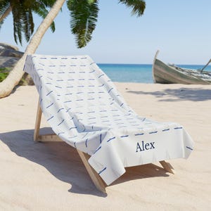 May include: A white beach towel with a blue sailboat pattern and the name "Alex" embroidered on it. The towel is draped over a wooden beach chair on a sandy beach.