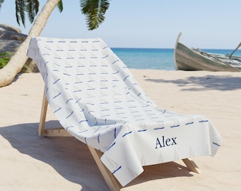 May include: A white beach towel with a blue sailboat pattern and the name "Alex" embroidered on it. The towel is draped over a wooden beach chair on a sandy beach.
