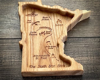 Puede incluir: Una talla de madera del estado de Minnesota con varias ciudades y lagos etiquetados. La talla es de color marrón claro y tiene una veta de madera natural. El texto "The North Star State" está tallado en la parte inferior de la talla.