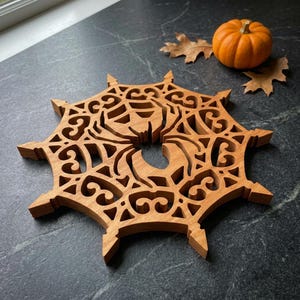 May include: A laser-cut wooden trivet featuring an intricate spider web design with a central spider silhouette. The light brown wood piece has a circular shape with pointed edges and decorative cutouts throughout the surface.