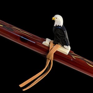 May include: A dark reddish-brown wooden flute featuring a carved bald eagle totem attached to the top with a light tan leather cord. The instrument has a smooth, polished finish with decorative yellow and black painted accents along the body.