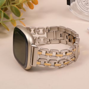 Könnte beinhalten: Ein silbernes und goldfarbenes Metall-Uhrenarmband. Das Armband hat ein Kettenglied-Design mit abwechselnd silbernen und goldenen Gliedern. Das Zifferblatt der Uhr ist quadratisch mit einem dunklen Bildschirm.