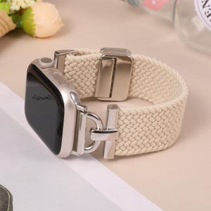Puede incluir: Correa de Apple Watch color crema con textura tejida. La correa presenta un cierre y una caja de reloj de color plateado. La esfera del reloj es negra. La correa está diseñada para un reloj inteligente.