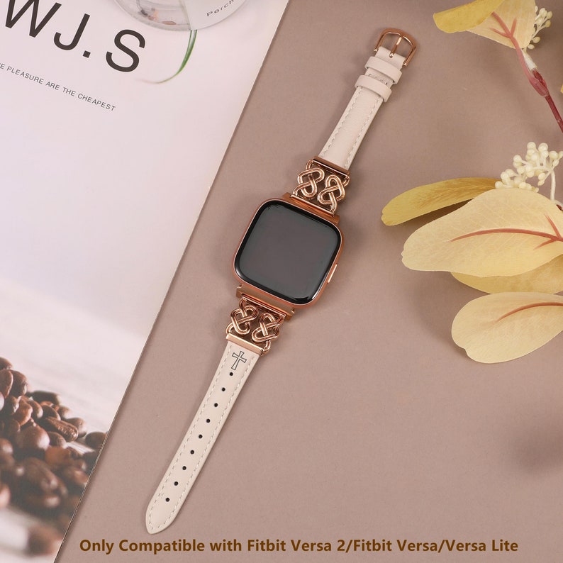 May include: A white leather watch band with a rose gold metal clasp and a cross design. The band is compatible with Fitbit Versa 2, Fitbit Versa, and Versa Lite.