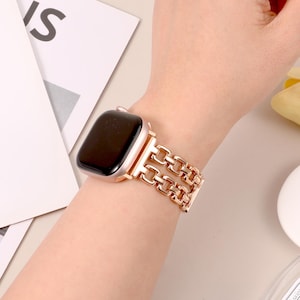 May include: A rose gold metal watch band with a linked chain design. The band is worn on a wrist.