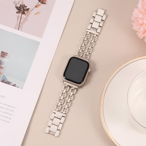 May include: A silver metal Apple Watch band with a double row chain link design. The band is shown on a white surface.