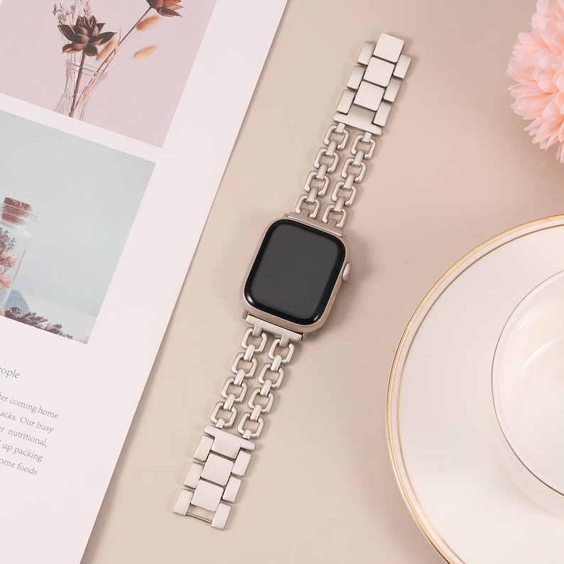 May include: A silver metal Apple Watch band with a double row chain link design. The band is shown on a white surface.