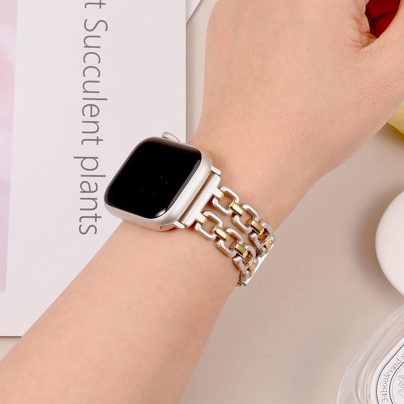 May include: A silver and gold chain link Apple Watch band. The band is made of two rows of chain links, with the top row being gold and the bottom row being silver.