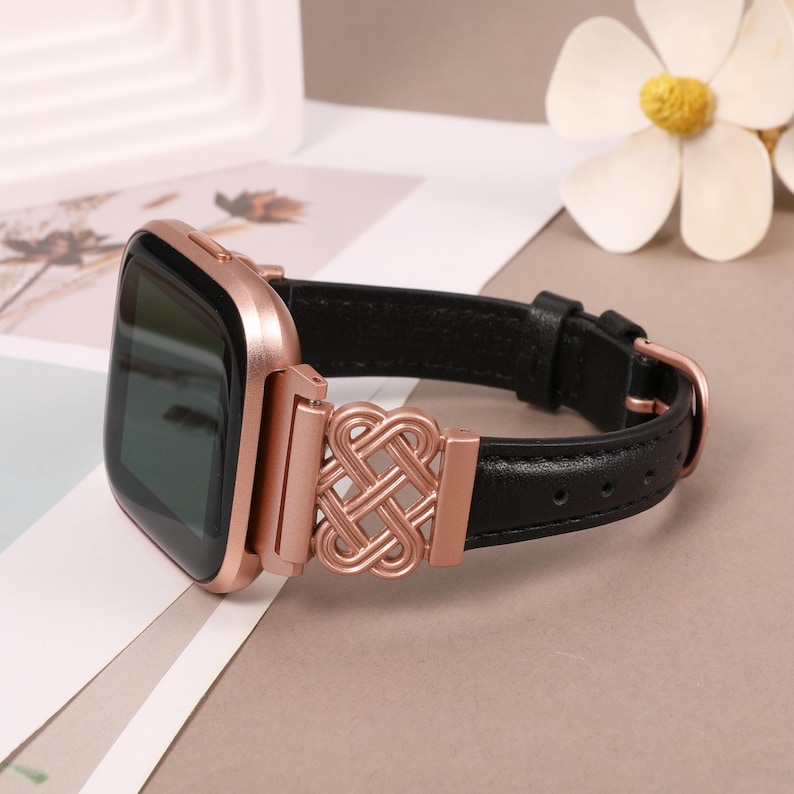 Puede incluir: Una correa de reloj de oro rosa con una correa de cuero negro y un cierre met&aacute;lico decorativo. El cierre presenta un dise&ntilde;o intrincado de c&iacute;rculos entrelazados.