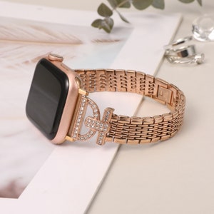 Puede incluir: Una pulsera de Apple Watch de oro rosa con un cierre engastado con diamantes. La pulsera está hecha de malla metálica y tiene un acabado pulido.