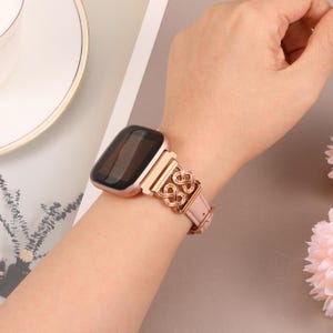 May include: A rose gold smartwatch with a rose gold metal band featuring a linked heart design. The band is attached to a square-shaped smartwatch with a black screen.