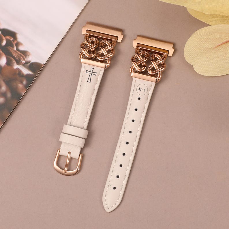 May include: Two white leather watch bands with rose gold metal clasps. One band has a cross design and the other has an interlocking circle design. Both bands have the letters "M-A" engraved on them.