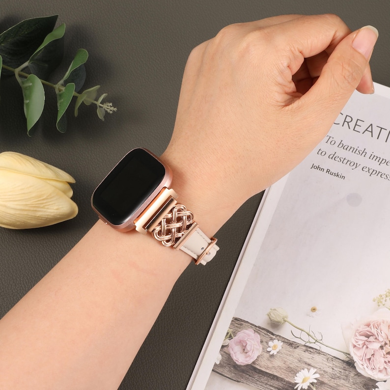 Puede incluir: Un reloj inteligente de oro rosa con una correa de cuero blanco con un dise&ntilde;o de metal trenzado. El reloj est&aacute; en la mu&ntilde;eca de una persona.