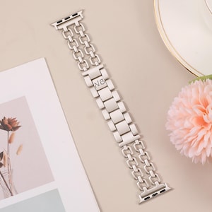May include: Silver metal Apple Watch band with a double row chain design and engraved initials "NB".
