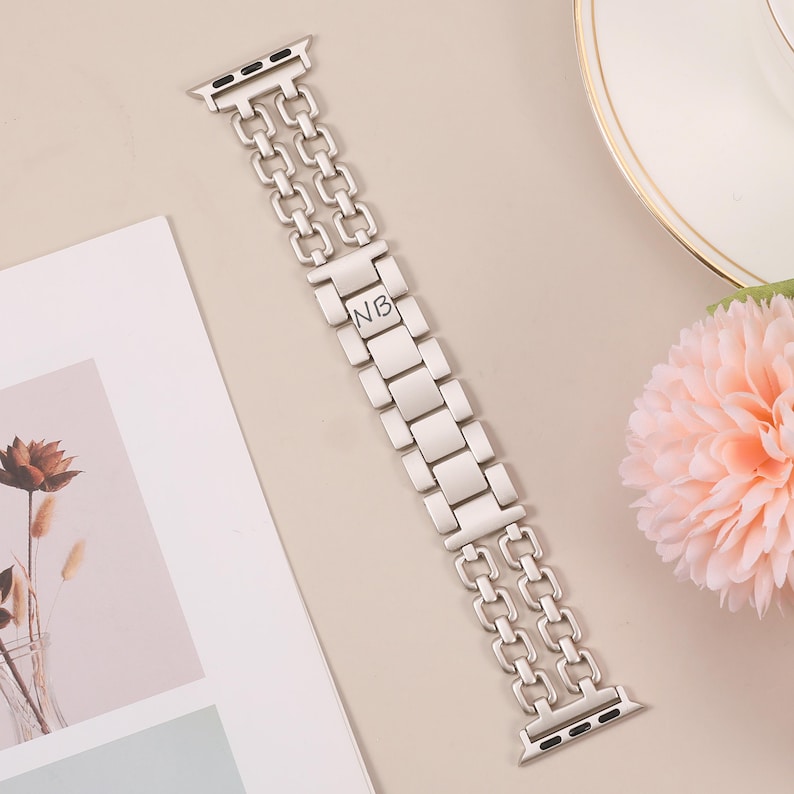 May include: Silver metal Apple Watch band with a double row chain design and engraved initials "NB".