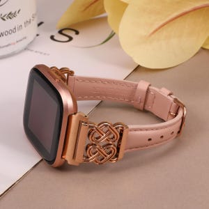May include: A rose gold smartwatch band with a pink leather strap and a rose gold metal clasp. The clasp features an intricate, interwoven design.