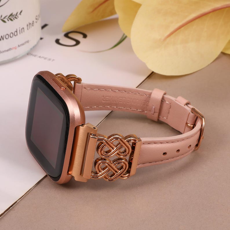 May include: A rose gold smartwatch band with a pink leather strap and a rose gold metal clasp. The clasp features an intricate, interwoven design.
