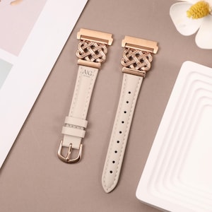 Könnte beinhalten: Zwei weiße Lederarmbänder mit roségoldenen Verschlüssen und einem dekorativen Knotendesign. Die Verschlüsse sind mit dem Text "AIG TOUSHI" graviert.
