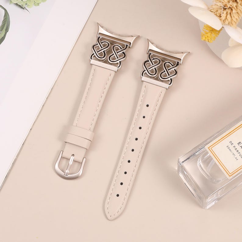 Pode incluir: Duas pulseiras de rel&oacute;gio de couro bege com fechos de metal prateado e designs decorativos de n&oacute;s.