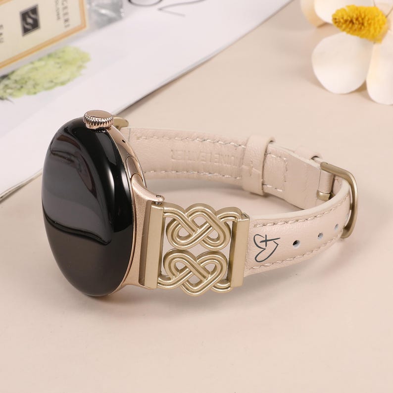 Pode incluir: Uma pulseira de rel&oacute;gio de couro creme com um fecho de metal dourado que apresenta um design de n&oacute; intrincado. A pulseira tem um design em forma de cora&ccedil;&atilde;o com uma letra 'K' dentro.