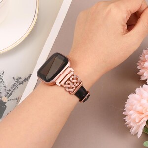 May include: A rose gold metal watch band with a black leather strap and a decorative knot design. The band is worn on a person's wrist.