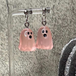 May include: A pair of pink ghost earrings. Each earring features a small, translucent ghost shape with black eyes and a white mouth. The earrings hang from silver-colored hooks and are displayed against a textured gray background.
