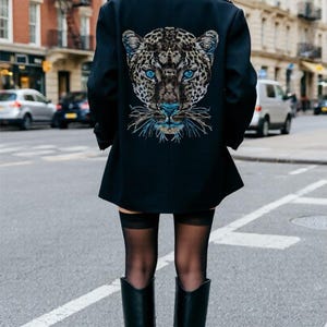 Könnte beinhalten: Schwarzer Mantel mit einem Leoparden-Gesicht-Design auf dem Rücken, mit blauen Augen und Akzenten. Der Mantel wird mit einer schwarzen Mütze, Strümpfen und kniehohen Stiefeln getragen. Das Bild wurde auf einer Straße aufgenommen.
