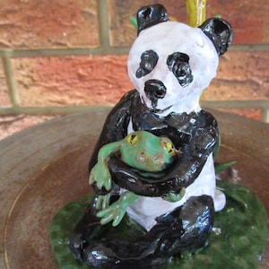May include: A ceramic panda figurine with black and white fur, holding a green frog figurine. The panda is sitting on a green base with brown accents.