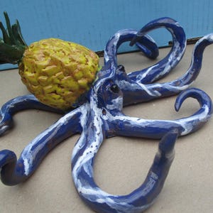 May include: A blue and white ceramic octopus with a yellow pineapple on its head. The octopus has eight tentacles and is painted with white swirls.
