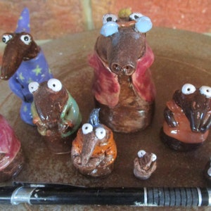 May include: A group of quirky ceramic figurines, each featuring a long-nosed character with large eyes. The figures are dressed in different outfits, such as a wizard's robe and a chef's coat. A pen is included for scale to show the size of the figurines.