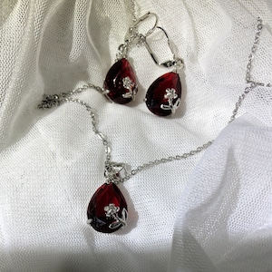 Puede incluir: Un conjunto de joyas que incluye un collar y pendientes. El conjunto presenta un collar de cadena plateada con un colgante en forma de lágrima de color rojo intenso y pendientes a juego. Cada pieza tiene un pequeño adorno floral plateado. Las joyas se exhiben sobre tela blanca texturizada.