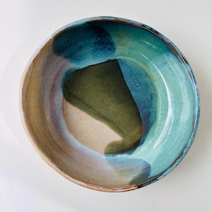 May include: A round ceramic bowl featuring a multi-coloured glaze with blended tones of deep teal, forest green, and sandy beige. The interior surface displays a smooth, glossy finish with organic colour transitions and a speckled texture.