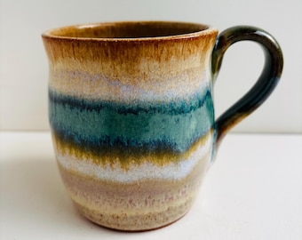 Op de afbeelding: Een keramische mok met een veelkleurig glazuur met horizontale banden in beige, wit, groenblauw en donkerblauw. De mok heeft een afgerond lichaam en een gebogen handvat, met een gladde, glanzende afwerking die vloeiend in elkaar overloopt.