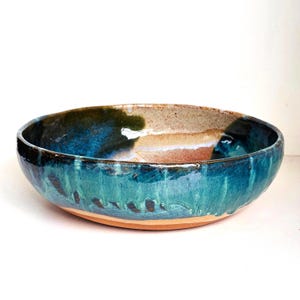 May include: A round ceramic bowl featuring a glossy glaze with deep blue, teal, and green tones that drip down the sides. The interior reveals a speckled beige and tan base, contrasting with the dark, vibrant exterior colours and smooth finish.