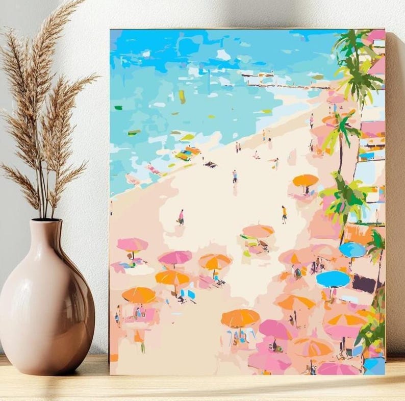 May include: A colorful beach scene painting with a light blue sky and turquoise water. The beach is filled with people, pink and orange umbrellas, and palm trees. The artwork is displayed on a wooden surface.