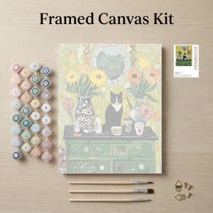 The image is a framed canvas kit featuring a painting of a cat sitting on a table surrounded by various objects, such as vases, cups, and a clock. The kit also includes paintbrushes, paint tubes, and a small picture frame.