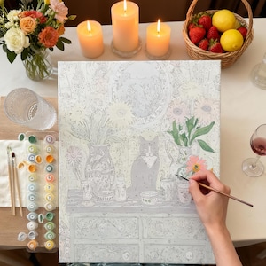 a person's hand painting a picture of a cat sitting on a table with various objects, such as vases, flowers, and a bowl. The painting is surrounded by art supplies, including paintbrushes and a palette of colors. There are also lit candles and a basket of fruit, including strawberries and lemons, in the background.