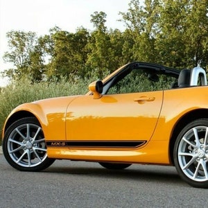May include: A yellow convertible sports car with black racing stripes and the text "MX-5" on the side. The car is parked on a tarmac surface with green trees in the background.
