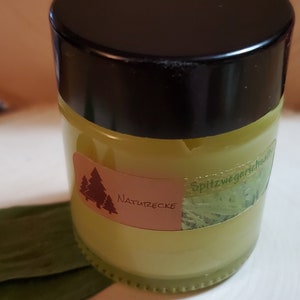 May include: A small glass jar with a black lid filled with a yellow cream. The jar has a label with the text "Naturzecke" and "Spitzwegerichhaltig" in brown lettering.
