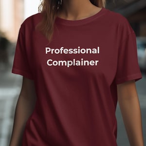 May include: A maroon t-shirt with the words "Professional Complainer" in white block letters. The shirt is a solid color and appears to be made of a soft material. The image is taken outdoors.