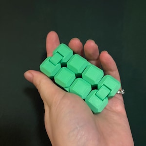 May include: A green fidget toy held in a hand. The toy is made of connected, cube-shaped pieces. The background is a solid, dark green color.