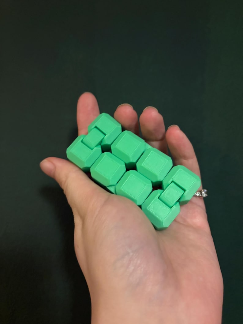 May include: A green fidget toy held in a hand. The toy is made of connected, cube-shaped pieces. The background is a solid, dark green color.
