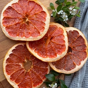 Può includere: Primo piano di quattro fette di pompelmo essiccate disposte su una superficie di legno. Le fette sono di un vibrante colore rosso-arancio con una buccia di colore chiaro. Piccoli fiori bianchi e foglie verdi sono sparsi intorno alla frutta.