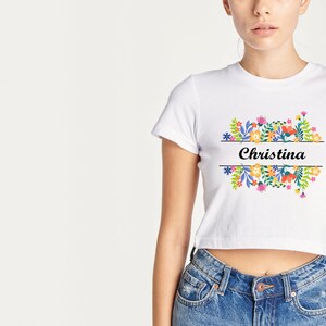 May include: White cropped t-shirt with a floral design and the name "Christina" printed on the front.