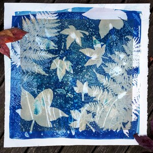 May include: A cyanotype print with leaf and fern silhouettes on a dark blue background. The leaves and ferns are in shades of white and grey, contrasting against the blue. The print is on white paper with a deckled edge.