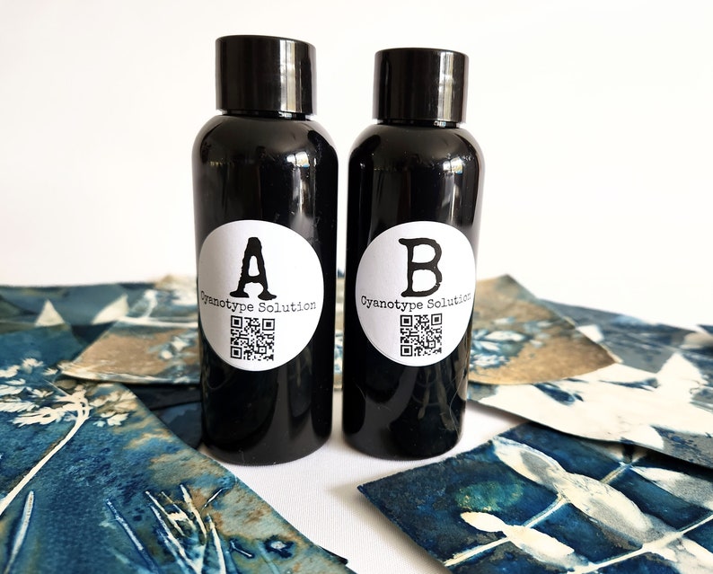 May include: Two black bottles, labelled "A" and "B", containing "Cyanotype Solution". The bottles have white labels and QR codes. They are placed on a white surface with blue and white cyanotype prints.