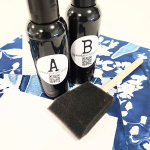 May include: Two black bottles marked "A" and "B" with "Cyanotype Solution" on white labels, a black foam brush, and blue and white patterned paper. The bottles contain a dark liquid, possibly a chemical solution for cyanotype printing.
