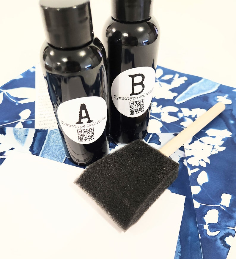 May include: Two black bottles marked "A" and "B" with "Cyanotype Solution" on white labels, a black foam brush, and blue and white patterned paper. The bottles contain a dark liquid, possibly a chemical solution for cyanotype printing.